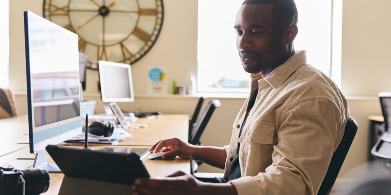Young creative black man working on computer in modern office at desk on tablet computer smiling